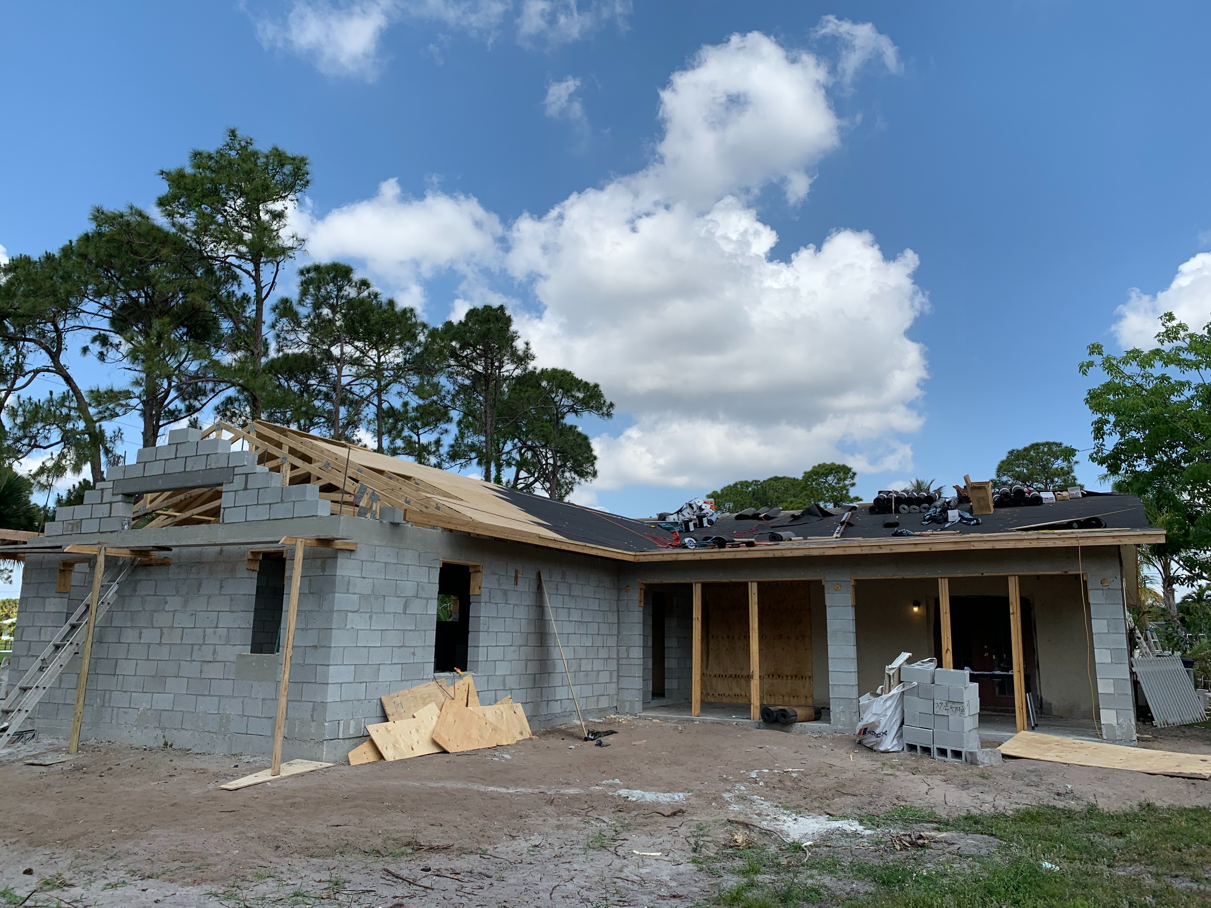 A house under construction with cinder block walls and a partially completed roof structure. Building materials and tools are placed on top of the roof, and plywood sheets are stacked in front of the building. The area surrounding the construction site is mostly dirt, with scattered building supplies. Tall pine trees and a partly cloudy sky are visible in the background.