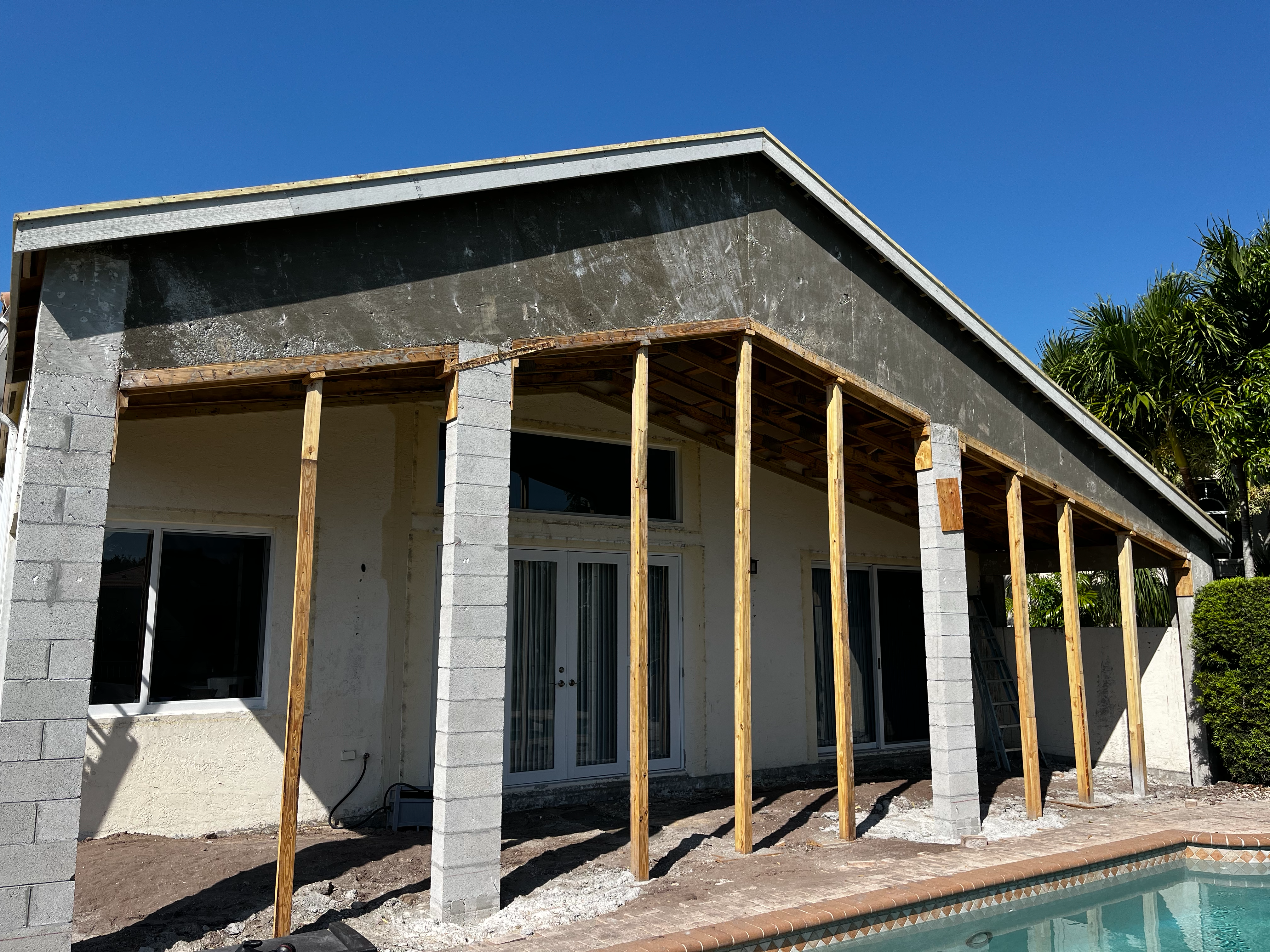 A partially constructed patio with wooden beams and cinder block columns in front of a house. The roof structure is in place, but the walls and exterior finishes are not yet completed. There is a pool in the foreground, and palm trees are visible in the background under a clear blue sky.