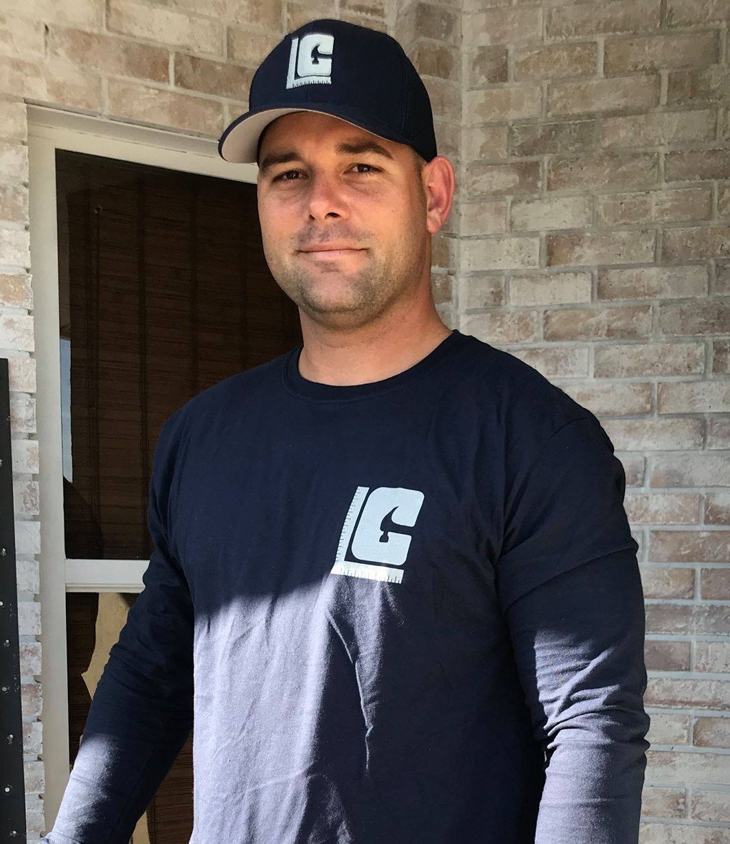 man wearing a navy blue long-sleeve shirt and matching navy baseball cap with the logo stands in front of a brick wall. He has a slight smile and is looking directly at the camera.