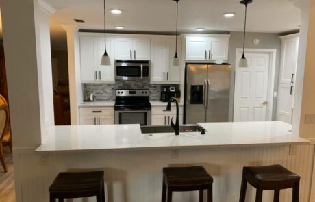 A modern kitchen with white cabinetry, stainless steel appliances, and a marble countertop island. Three pendant lights hang over the island, which has a built-in sink and is paired with three bar stools. The backsplash features a gray stone pattern, adding texture to the space.