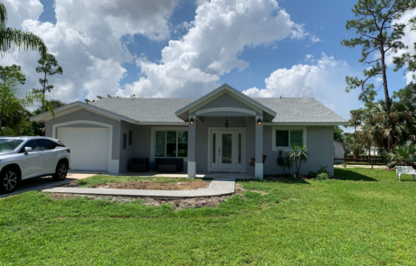 A finished single-story home with a light gray exterior and a simple front porch. The house has a two-car driveway with an SUV parked on the left side. There is a well-maintained green lawn in the foreground and palm trees in the background. The sky is bright with scattered clouds.