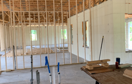 Interior view of a home under construction, showing exposed wooden roof trusses and unfinished wall framing. Plumbing pipes are visible in the concrete floor, and a worktable with tools and materials is set up nearby. The space is open and ready for further development, with natural light coming in through window openings.