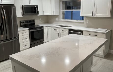 A contemporary kitchen with sleek white cabinets, stainless steel appliances, and a large quartz island countertop. Two pendant lights with geometric glass shades hang over the island, creating a warm ambiance. The space is illuminated by recessed ceiling lights, enhancing the clean and modern design.