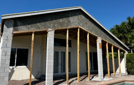 A partially constructed patio with wooden beams and cinder block columns in front of a house. The roof structure is in place, but the walls and exterior finishes are not yet completed. There is a pool in the foreground, and palm trees are visible in the background under a clear blue sky.