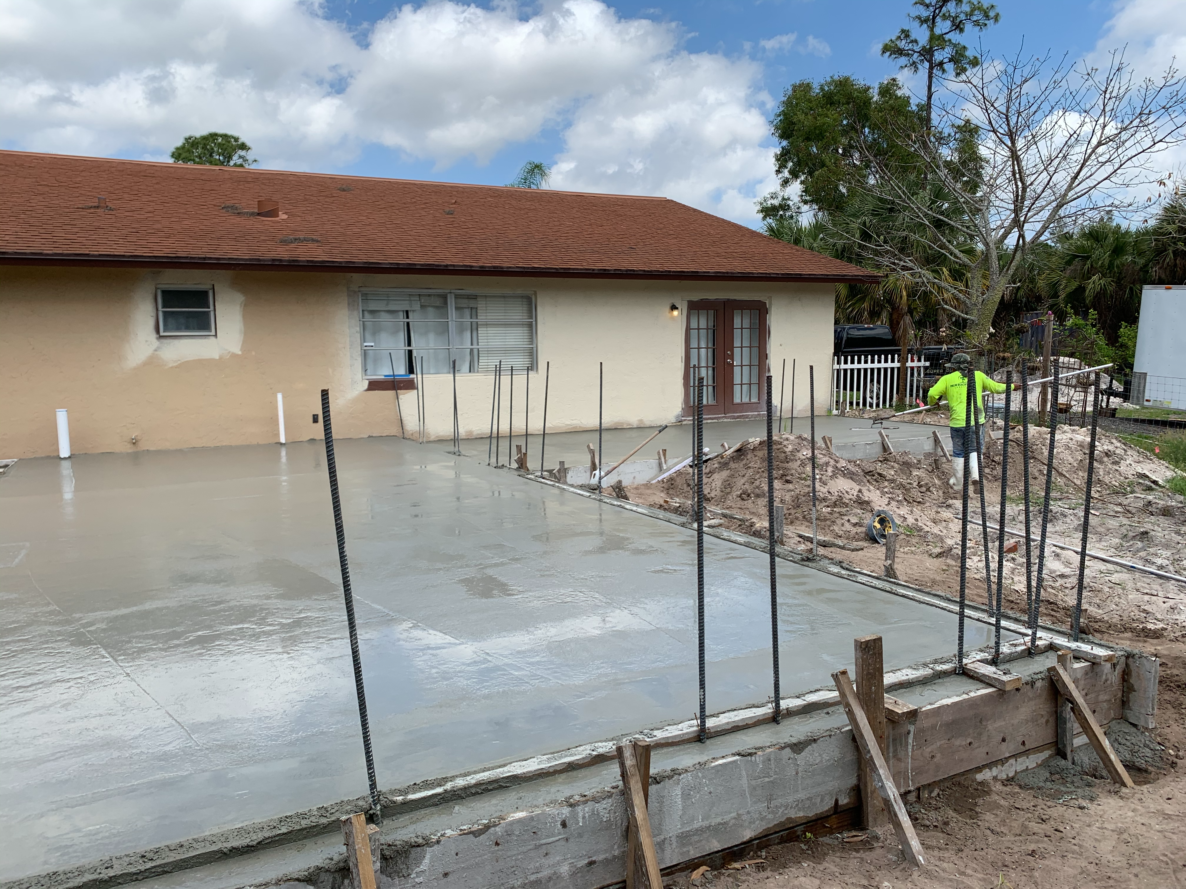 A construction site with freshly poured concrete forming the foundation for an addition to an existing house. Rebar rods are positioned upright along the edges of the slab for future wall support. A worker in a green shirt is seen in the background near a mound of dirt, preparing the site for the next phase. The existing house has a beige exterior with a red shingle roof and French doors.